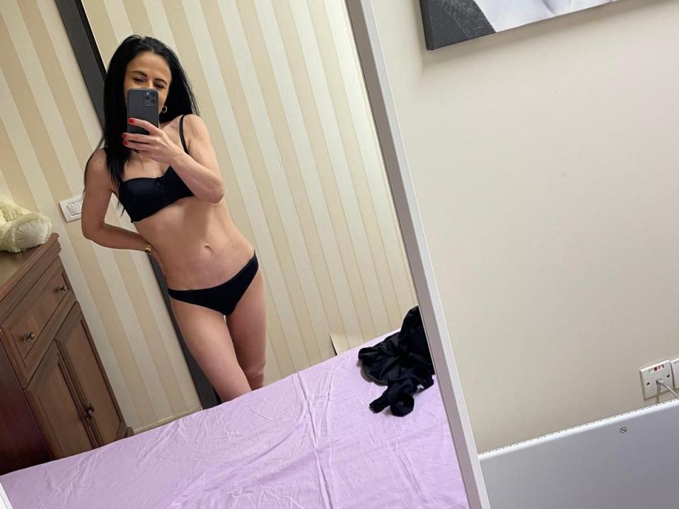 BrianaFlynt with black bra and panties, standing in front of mirror, holding phone, showing her tits and ass, with a pink bedsheet and black clothes on it.