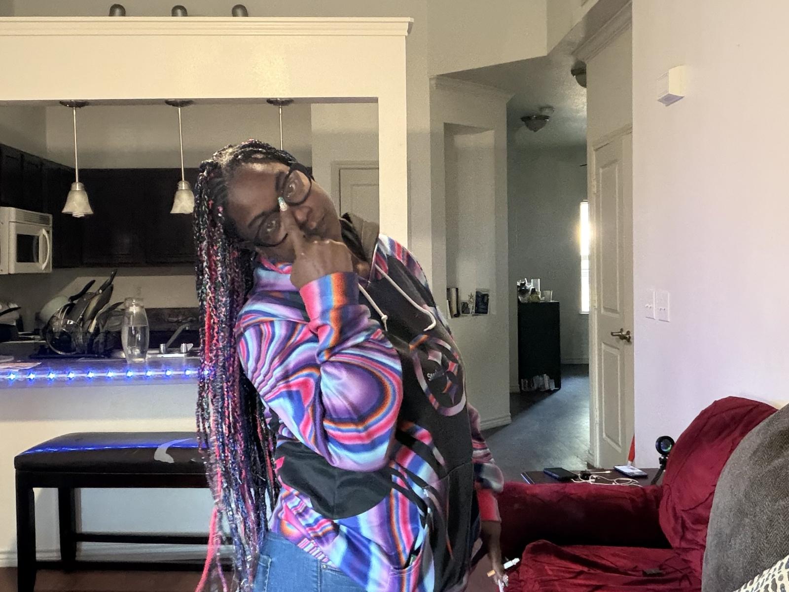 WetVibez with dark skin, long red and pink braids, wearing glasses, colorful patterned jacket, covering her small tits with one hand, standing in a modern kitchen.