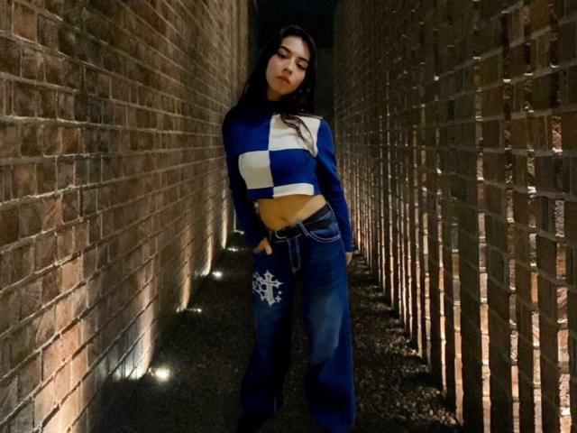 KatherinJohns with long black hair, wearing a blue and white striped crop top, exposing her taut abs, and loose blue jeans with a snowflake design, standing in a brick alley.