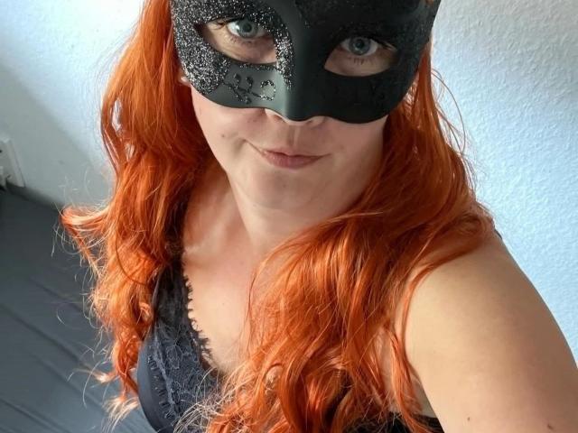 SarahFoxx-hot with orange hair, wearing a black glitter mask, black lace top, smiling at the camera, showing her blue eyes, and a cute face, in a selfie.