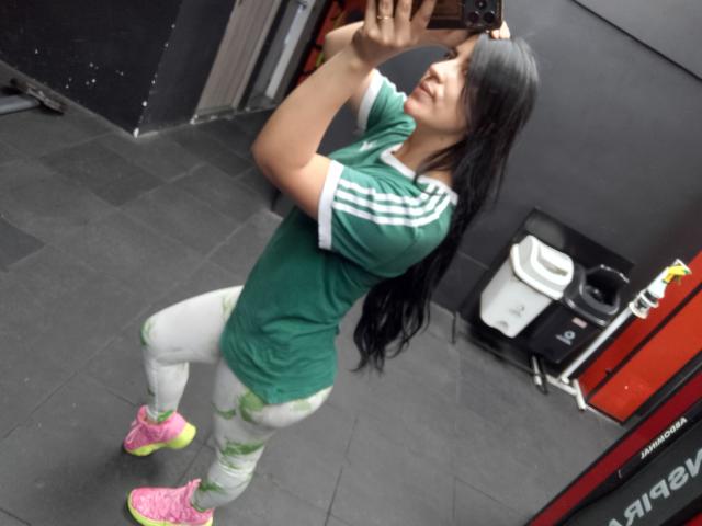 SofiaFountaine with long black hair, green shirt, white leggings with green patterns, pink sneakers, holding phone, taking mirror selfie in gym, showing her ass and small tits.