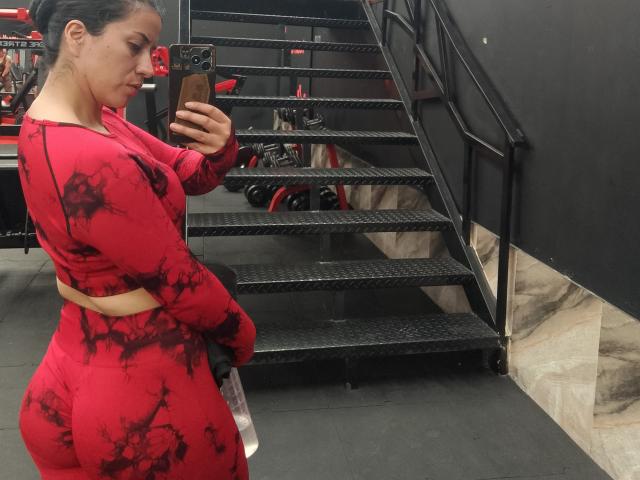 SofiaFountaine with a curvy ass, wearing a red and black tie-dye sports outfit, holding a phone, taking a mirror selfie in a gym with metal stairs.
