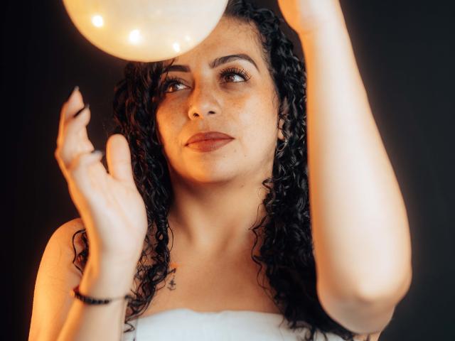 Alissa, con cabello negro rizado y pecas, sosteniendo una esfera brillante cerca de su rostro, mirando hacia arriba con una ligera sonrisa, vestida con un top blanco sin tirantes..