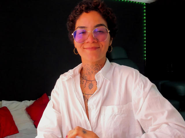 AnaRoa, met krullend bruin haar, draagt roze zonnebrillen en een witte blouse met knopen, en toont tatoeages op haar nek en borst. Ze glimlacht en draagt gouden oorringen..