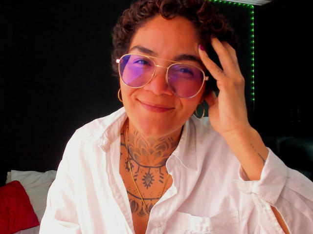AnaRoa with curly brown hair, wearing pink reflective sunglasses, a white shirt, and showing tattooed neck, smiling, touching her head, with purple nail polish.