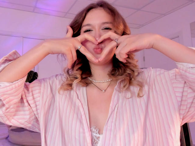 Synrix with wavy blonde hair, pink striped shirt, and pearl necklace, making a heart shape with her hands and sticking out her tongue. Her tits are slightly visible.