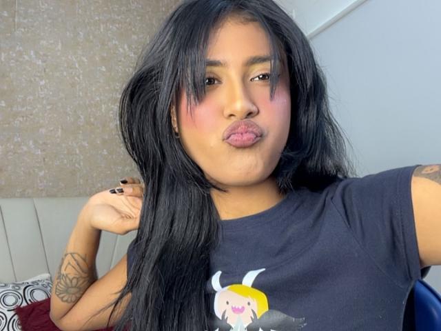 Babilett heeft lang zwart haar, volle roze lippen en zwarte nagels, draagt een zwart shirt met een cartoon erop, heeft tatoeages op haar armen en maakt een fronsende gezicht..