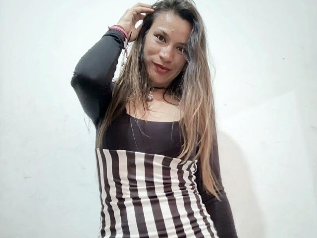 AlannaFox with long brown hair, wearing a black and white striped dress, black long sleeves, and red lipstick, smiling with hand in hair. She's got a cute face and small tits.