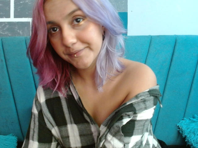 AlterEgoSecond with pink and purple hair, smiling, wearing an off-shoulder plaid shirt, showing her tits, has a lip piercing, sitting on a blue upholstered seat.