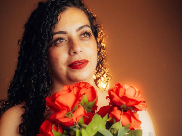 Alissa, con i capelli neri ricci, rossetto rosso, che tiene rose rosse vivaci, luce soffusa, sfondo marrone caldo, guarda verso l'alto con un leggero sorriso, bellissima, sexy, romantica, delicata..
