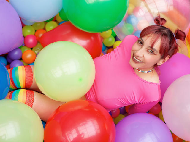AliRousse com cabelo ruivo, vestindo uma blusa rosa, meias coloridas listradas, cercada por balões, sorrindo, cheinha, fofa, pele clara, usando um colar, deitada em uma piscina de balões coloridos..