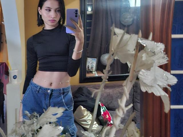 EllectraX, avec ses courts cheveux noirs, portant un débardeur noir à manches longues et un jean bleu, prend un selfie devant un miroir, montrant un corps mince et une silhouette plate, avec une chaîne à la taille et des fleurs..