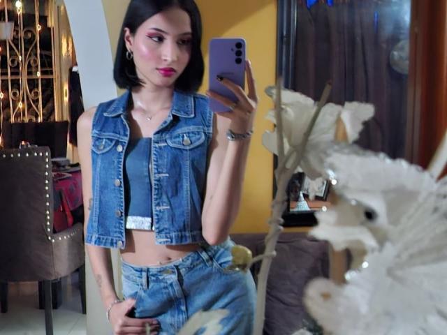 EllectraX, avec ses courts cheveux noirs, portant un gilet en jean et un jean taille haute, tenant un téléphone violet, montrant un tatouage sur sa main droite, se tient dans une pièce..