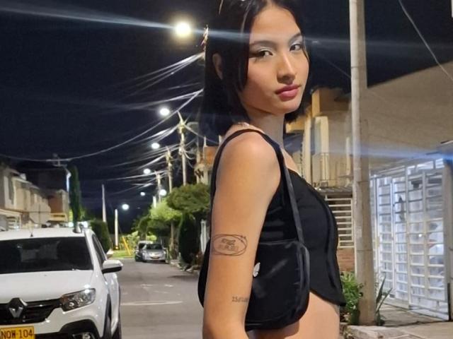 EllectraX has a cute face, dark straight hair, black crop top, small tits, tattoo on right arm, standing on street at night, white car in background, looking over shoulder.