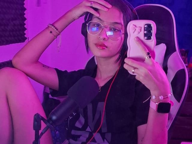 EllectraX, avec ses courts cheveux noirs, ses lunettes, son anneau de nez et son rouge à lèvres rose, est assise sur une chaise, tenant un téléphone, portant une chemise noire et un casque, devant un microphone..