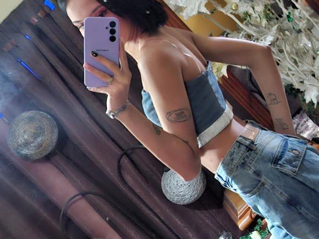 EllectraX, avec ses courts cheveux noirs, portant un haut moulant gris et un jean bleu, montre ses tatouages sur ses bras, tenant un iPhone violet, dont le reflet apparaît dans un miroir..