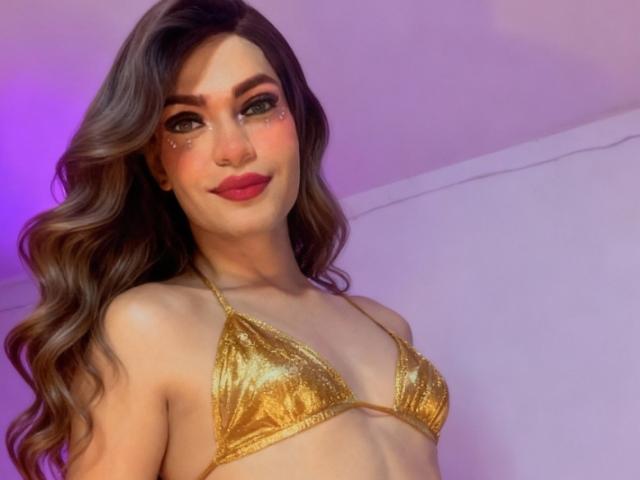 GingerSexyX with long wavy brown hair, wearing a shiny gold bikini top, showing small tits, and a cute nose ring, smiling with pink lipstick.