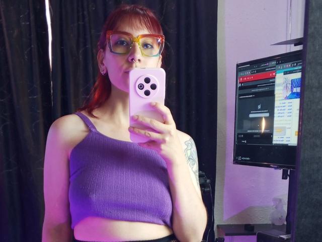 AliRousse with red hair, glasses, and a purple ribbed crop top, holding a pink phone, showing a tattoo on her left arm, in a mirror selfie.