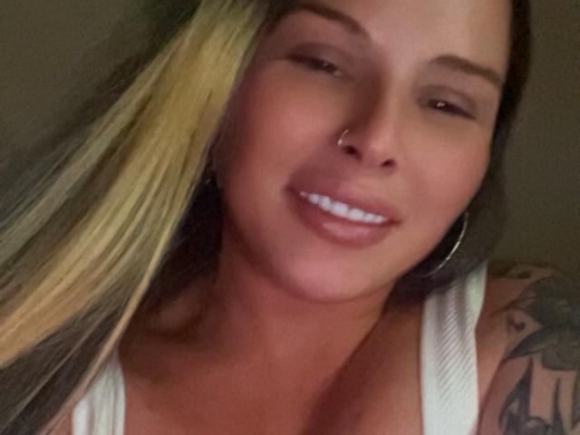 MonyBelle has blonde and brown hair, a nose ring, and a tattoo on her right shoulder. She's smiling with white teeth, wearing a white tank top.