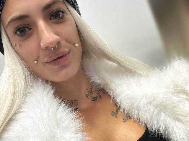 MrsSkinnyGoddess-hot with white hair, nose piercings, and facial piercings, wearing a fluffy white jacket, showing tattoos on her chest, and a black headband, smiling softly.