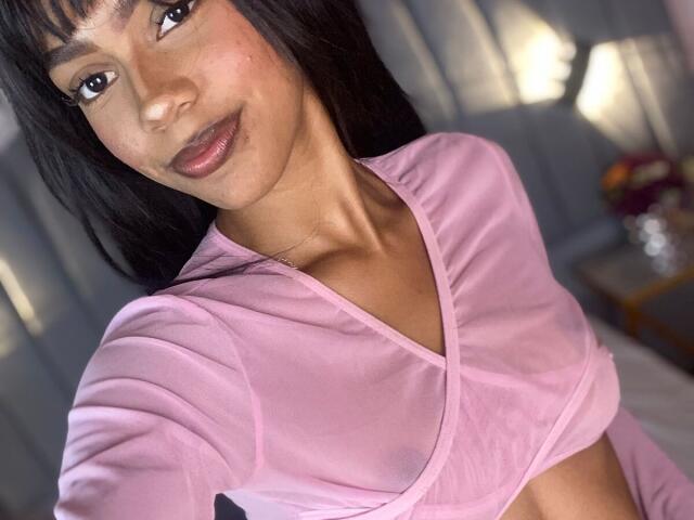 AylaCallowey with dark hair, brown eyes, and pink sheer top showing small tits, smiling at camera, light brown skin, subtle makeup, bedroom background, soft light.
