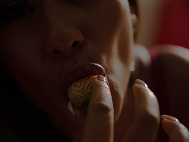 AlessandraMalory's face with glossy lips, gently sucking a strawberry held by her fingers. Her pinkish lips are slightly shiny, and her fingers are close to her mouth.