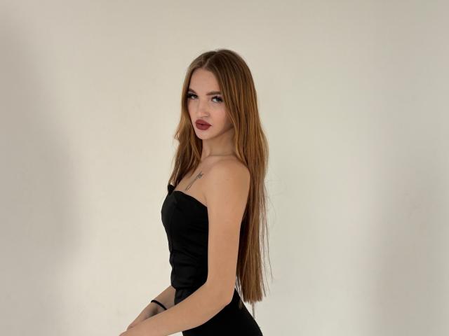 KlarnetHot with long brown hair, wearing a black strapless dress, showing her small tits and a tattoo on her right chest, looking at the camera with red lips.