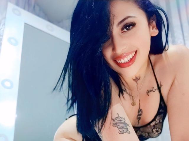 AngelDamons with black hair, smiling, showing tits in a lace bra, tattoos on chest and arm, nose ring, red lipstick, leaning forward, in a selfie.