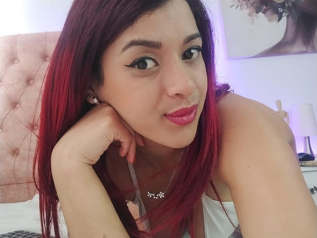 SweetieAlice69 with red hair, pink lipstick, and eyeliner, leaning on her hand, showing her tits in a pink strap dress, wearing a necklace, in a bedroom.