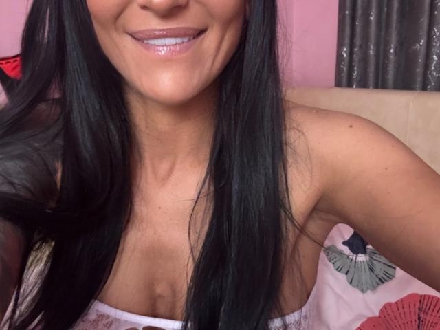AndraD with long black hair, shiny pink lips, smiling, showing cleavage, and tan skin, wearing a pink strapless top, in a bedroom.
