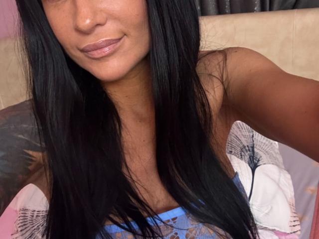AndraD with long black hair, brown skin, and pink lips, wearing a blue lace top, showing her tits, smiling slightly, in a bed with a patterned pillow.