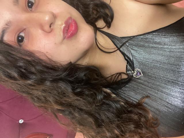 SharaSchart with curly brown hair, glossy pink lips, wearing a black shiny top with a heart necklace, looking at the camera, showing a bit of her chest.