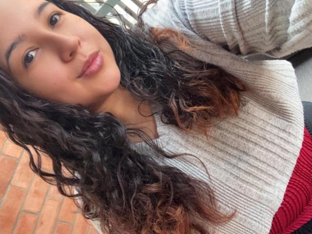 SharaSchart has curly brown hair, light pink lips, and a gray rib-knit sweater with red pants. Her cute face and small nose are visible. She's taking a selfie.