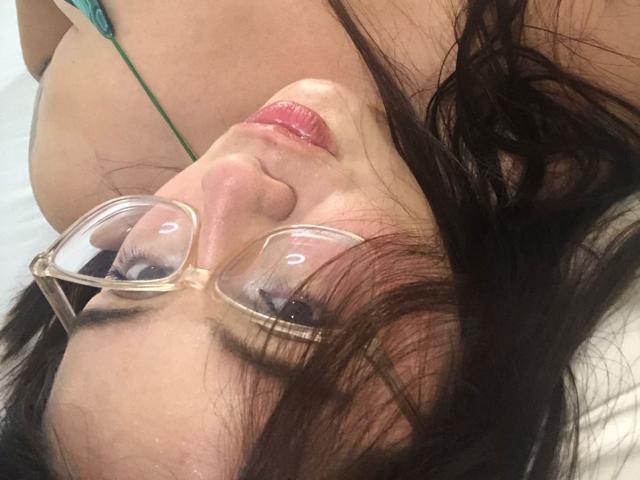 CaroliineRoss liegt mit Brille, dunklem Haar, pinkem Lippenstift und einer grünen Halskette da. Ihre Brüste sind im Hintergrund teilweise sichtbar. Ihre Nase ist leicht nach oben gebogen..
