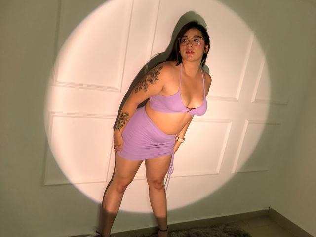 CaroliineRoss mit großen Brüsten, einem wohlgeformten Hintern und einem violetten Bikini-Oberteil und -Rock, steht im Scheinwerferlicht und zeigt Tattoos an ihrem linken Arm und ihrer rechten Schulter..