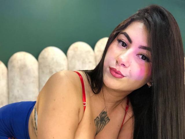 AlanaaWalker with pink and purple makeup, long brown hair, red bra, blue dress, tattoos on shoulder and chest, leaning forward, pouty lips, white picket fence background.