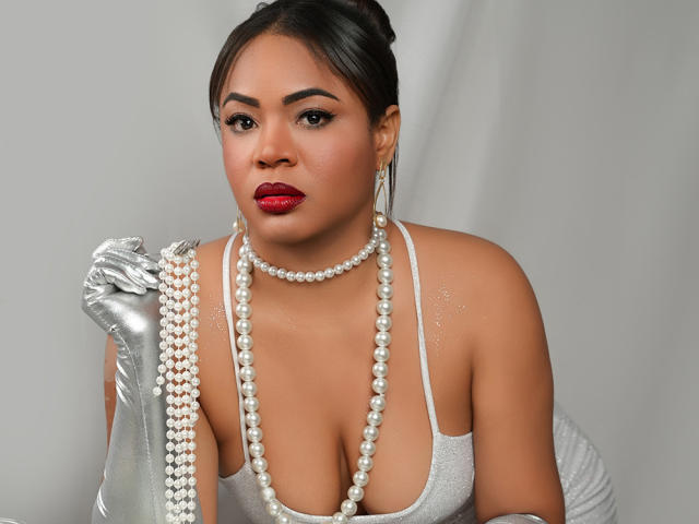 AliceBooty with red lips, wearing a silver glove, holding a pearl necklace, and a sparkly white dress, showing her large tits and a cute, confident face.