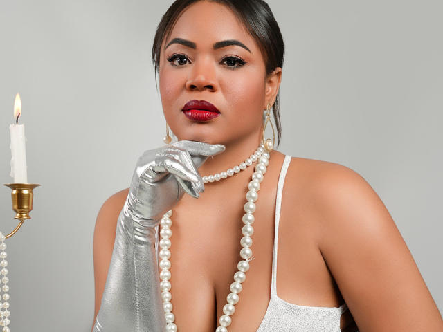 AliceBooty with red lipstick, shiny silver gloves, white pearl necklace, and earrings, holding a candle, wearing a low-cut, sparkly white dress, showing her tits.