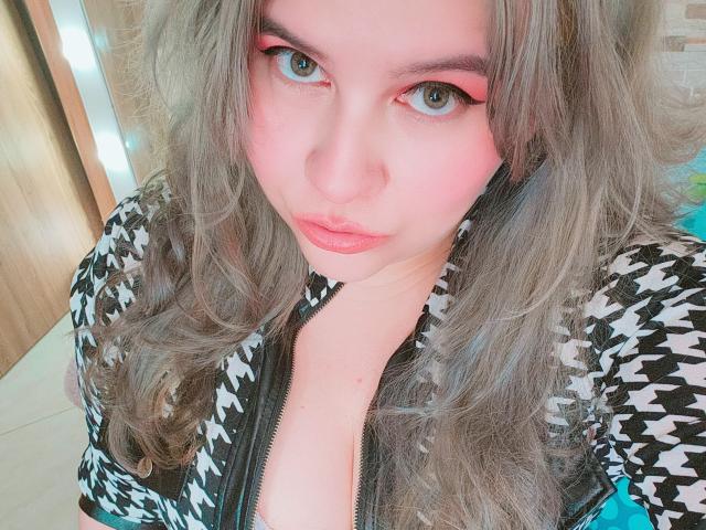 Akkanee with pink eyeshadow, wavy gray hair, pouty pink lips, black and white houndstooth jacket, blue zipper, and pale skin, looking directly at the camera.