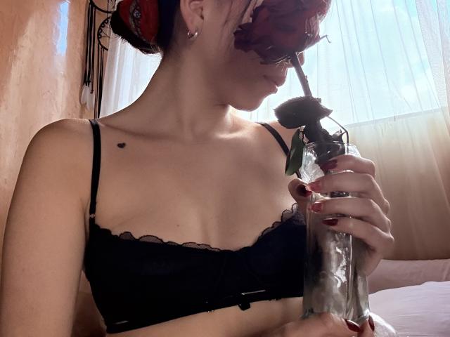 IzzysMisss with red hair, black lace bra, holding a clear glass with a black rose, sipping. She's got a small black heart tattoo near her right boob.