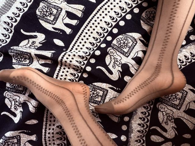 IzzysMisss' feet in sheer black stockings with back seam patterns, laying on a dark blue patterned fabric with white elephant and floral designs.