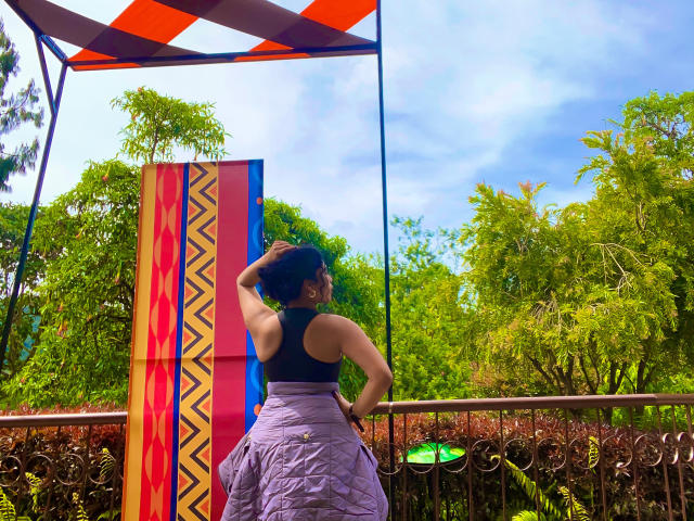 SophieSonyas with her back to the camera, black tank top, purple skirt, hand in hair, standing next to colorful vertical pattern, under orange canopy, with green trees.