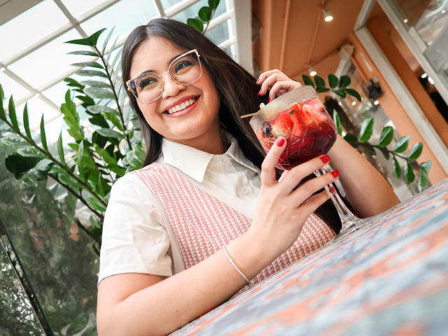 CamilaSofiia with glasses, smiling, holding a drink with fruit, white shirt, pink herringbone vest, red nails, silver bracelet, in a bright indoor setting, looking happy.