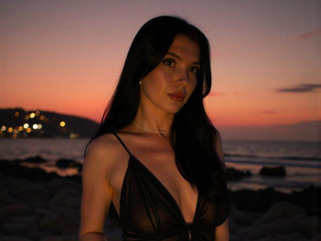 AngelSweetShy mit dunklem Haar, trägt ein durchsichtiges schwarzes Kleid, das ihre Brüste zeigt, steht auf einem felsigen Strand bei Sonnenuntergang, trägt eine goldene Halskette und schaut in die Kamera..