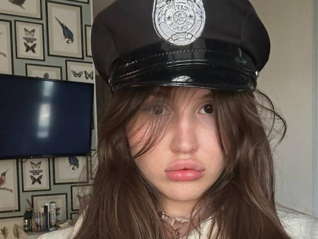 AnjelinaRos has long brown hair partially covering her face, wearing a black police hat with a shiny, reflective black brim and a silver police badge. She has fair skin, pinkish lips, and brown eyes. The hat casts a slight shadow over her face. The background is blurred, showing framed insect illustrations and a flat-screen TV. She wears a white collar with a small, metallic ring at the center. The image has a casual, slightly disheveled look with a focus on her facial expression and hat.