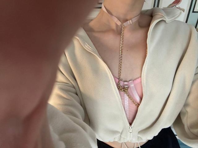 AnjelinaRos indossa una felpa con zip color crema, che lascia intravedere un reggiseno a righe rosa e bianche, con una catena dorata che pende e una collana rosa, in un selfie..