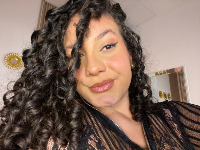 RachellBeaumont has curly black hair, full glossy lips, and is wearing a black lace top, smiling with a slight nose tilt, and winged eyeliner.