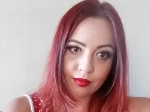 LadyHellena has red lipstick, red eyes, and red hair, looking directly at the camera with a slight smile, wearing a red polka dot top, and large hoop earrings.
