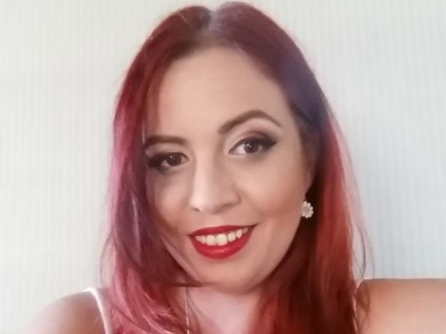 LadyHellena has red hair, red lipstick, and is smiling with her tits slightly visible in a white top. She's wearing a diamond earring and has dark eye makeup.