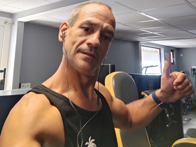 NousCoquins, bald with short gray hair, wearing a black tank top, earbuds, and a blue wristband, giving a thumbs-up, muscular, slightly smiling, gray office in background.